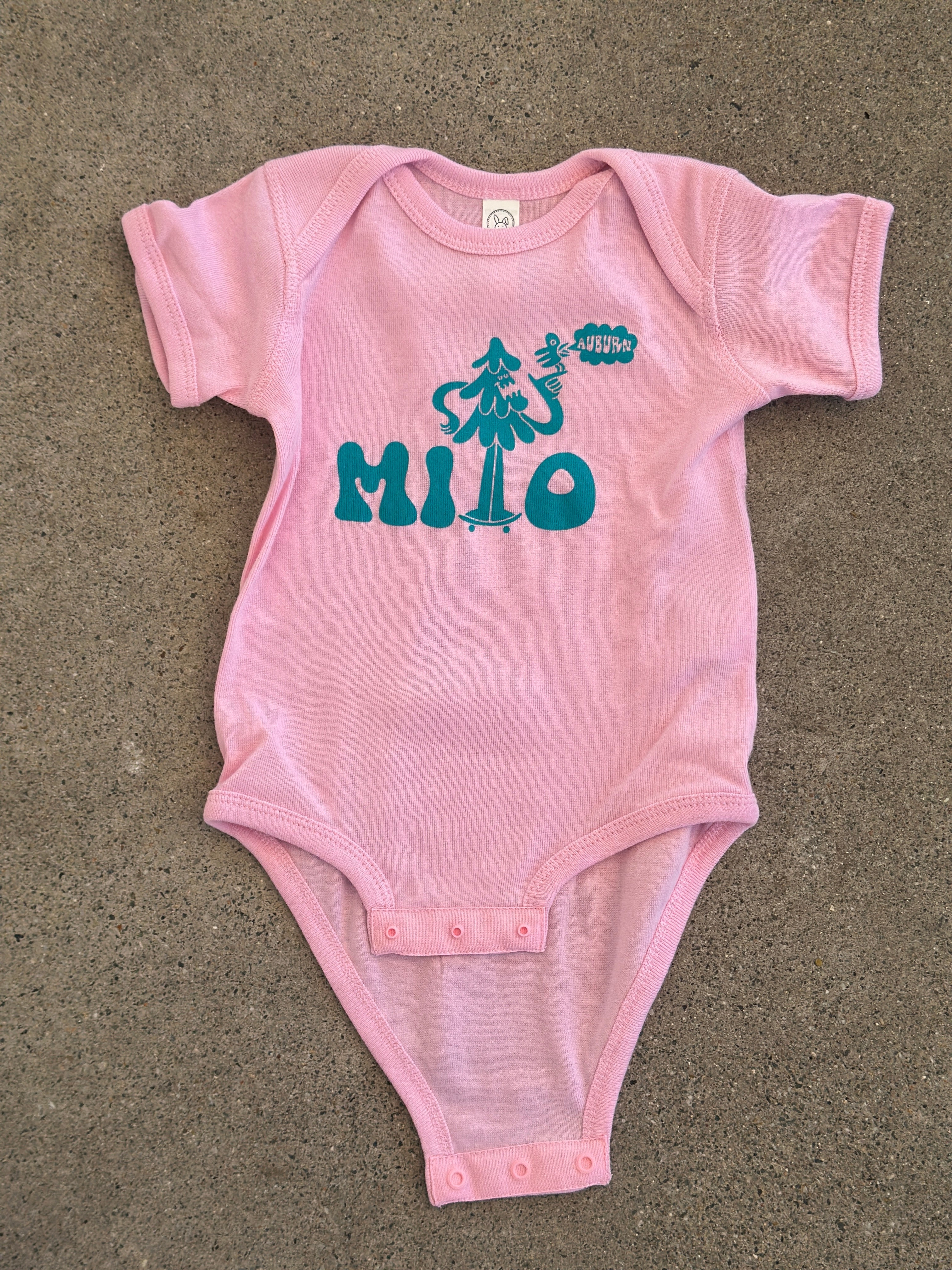 Pink baby onesie with teal text on a beige surface